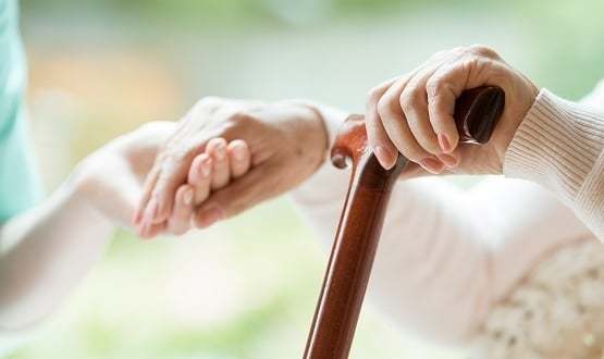 A care provider holds the hand of an elderly woman with a walking cane