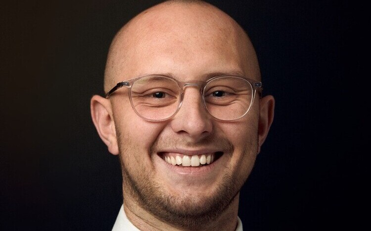 Jordan Fulcher, Clinical Solutions Consultant for EMEA, smiling in a professional headshot against a dark background, wearing glasses and business attire.